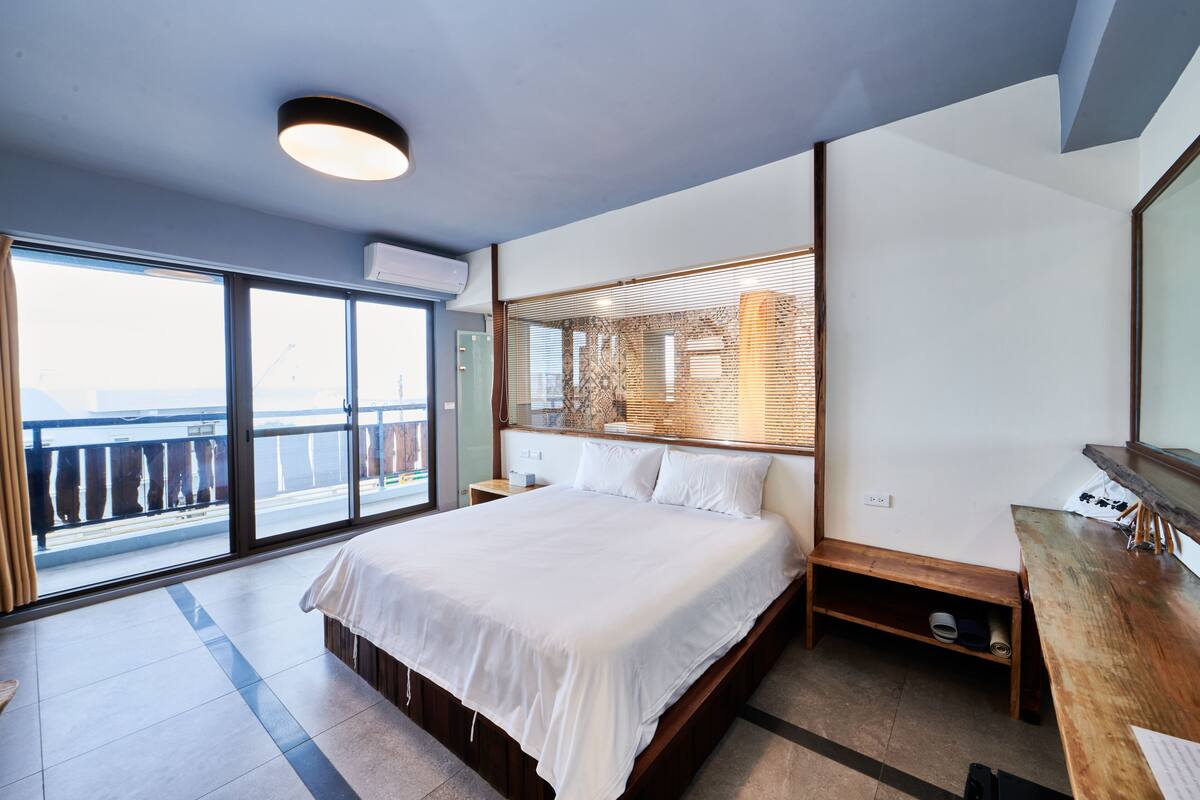 A modern bedroom with a large bed, a wooden nightstand, a desk, and a view of the outdoors through large windows.