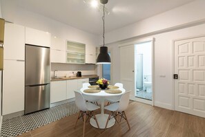 A modern kitchen with white cabinets, a stainless steel refrigerator, a round dining table with white chairs, and a black pendant light.