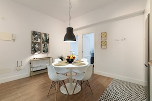 A dining area with a round table, white chairs, a black pendant light, and a fruit bowl.