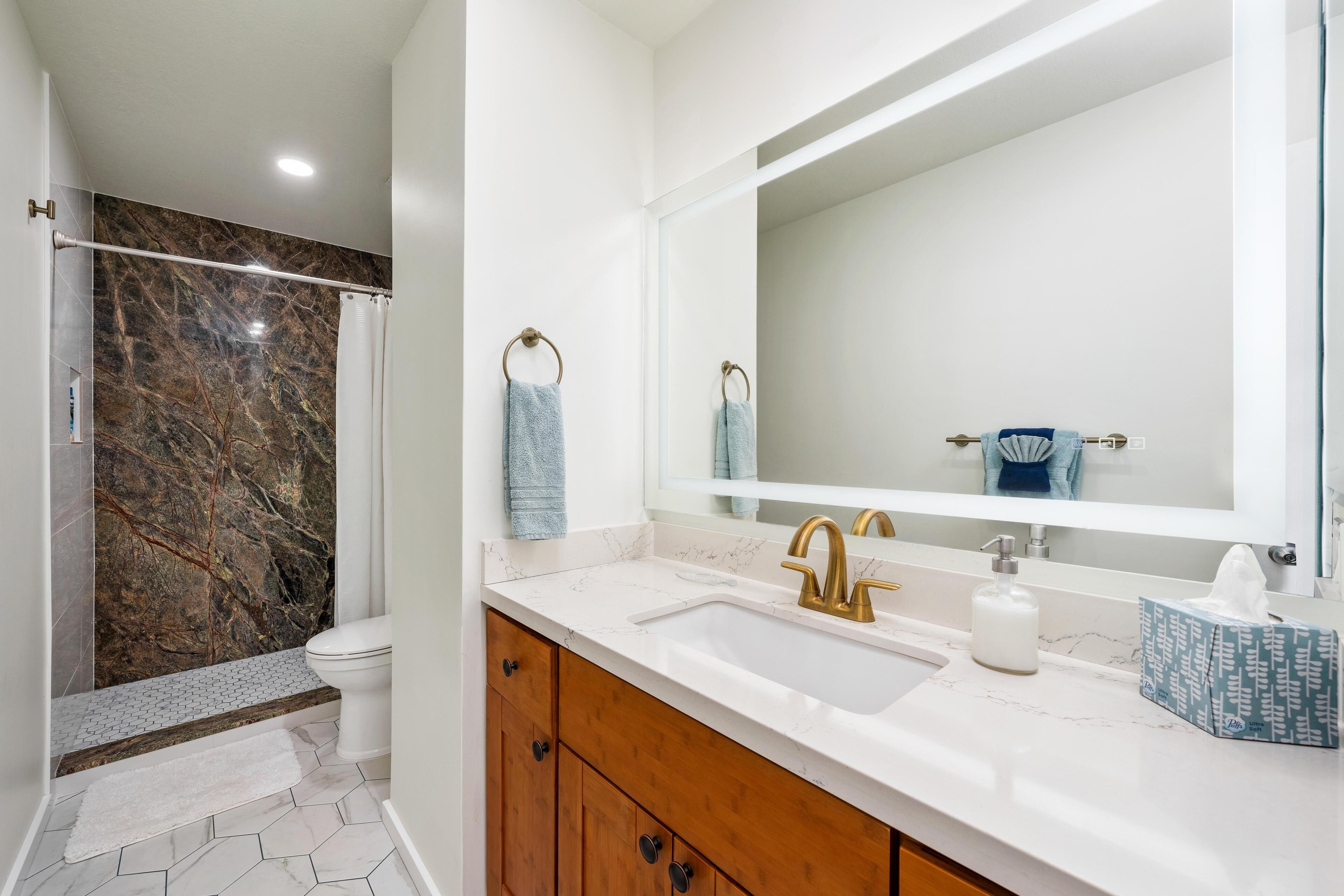 A modern bathroom with a marble shower, a white sink with gold fixtures, and a large mirror.