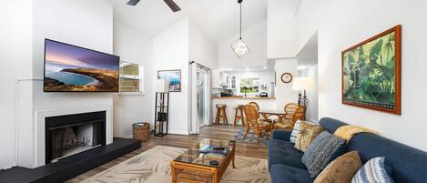 A spacious living room with a fireplace, a large flat-screen TV, a ceiling fan, and a dining area in the background.