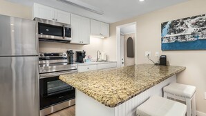 A modern kitchen with stainless steel appliances, a granite countertop, white cabinetry, and a painting of a coastal scene. - 1030 Second Street 1 Bedroom Home by RedAwning (Fort Myers Beach)
