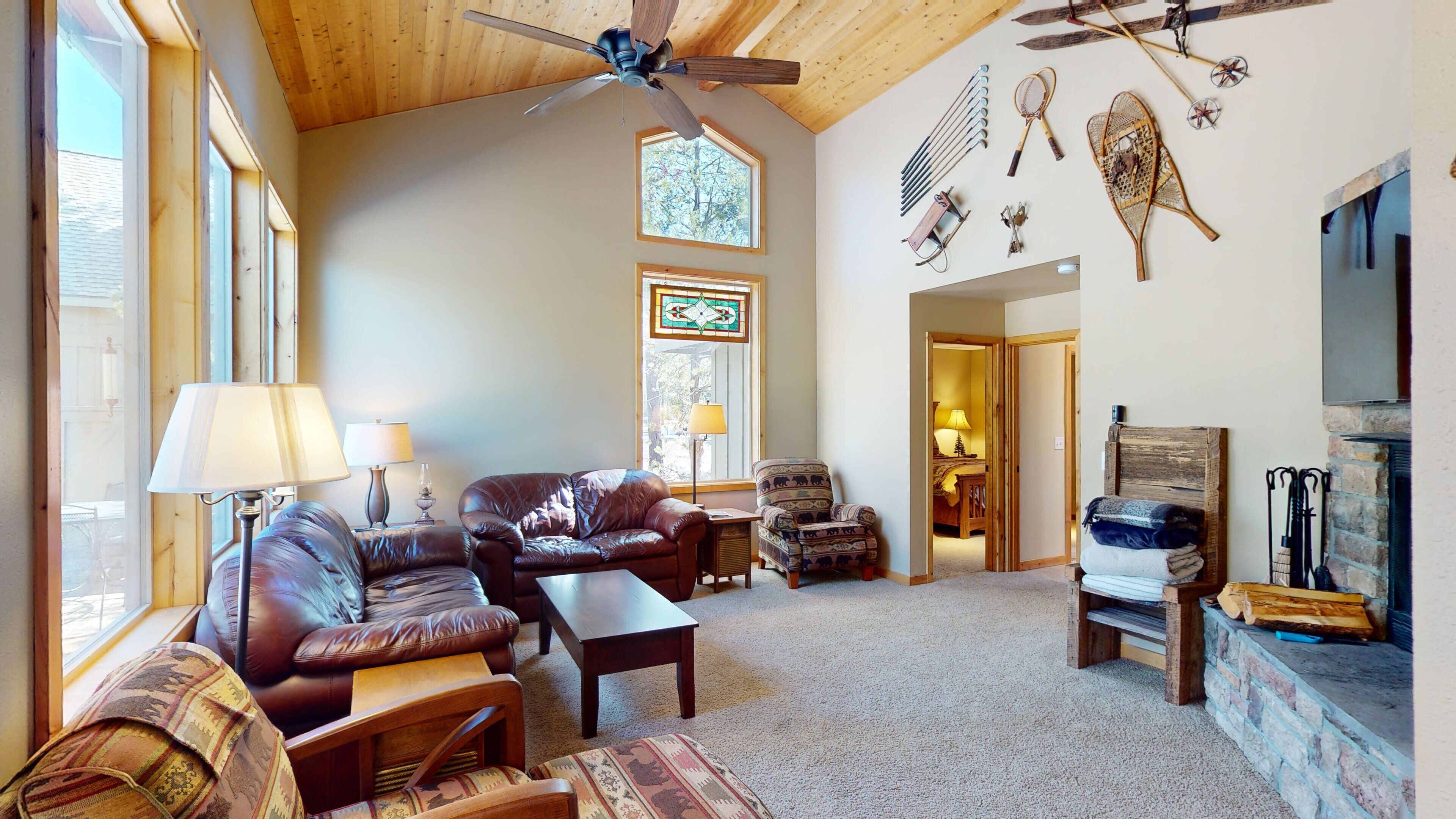 A spacious living room with leather sofas, a wooden coffee table, and a fireplace. There are multiple windows and a ceiling fan.
