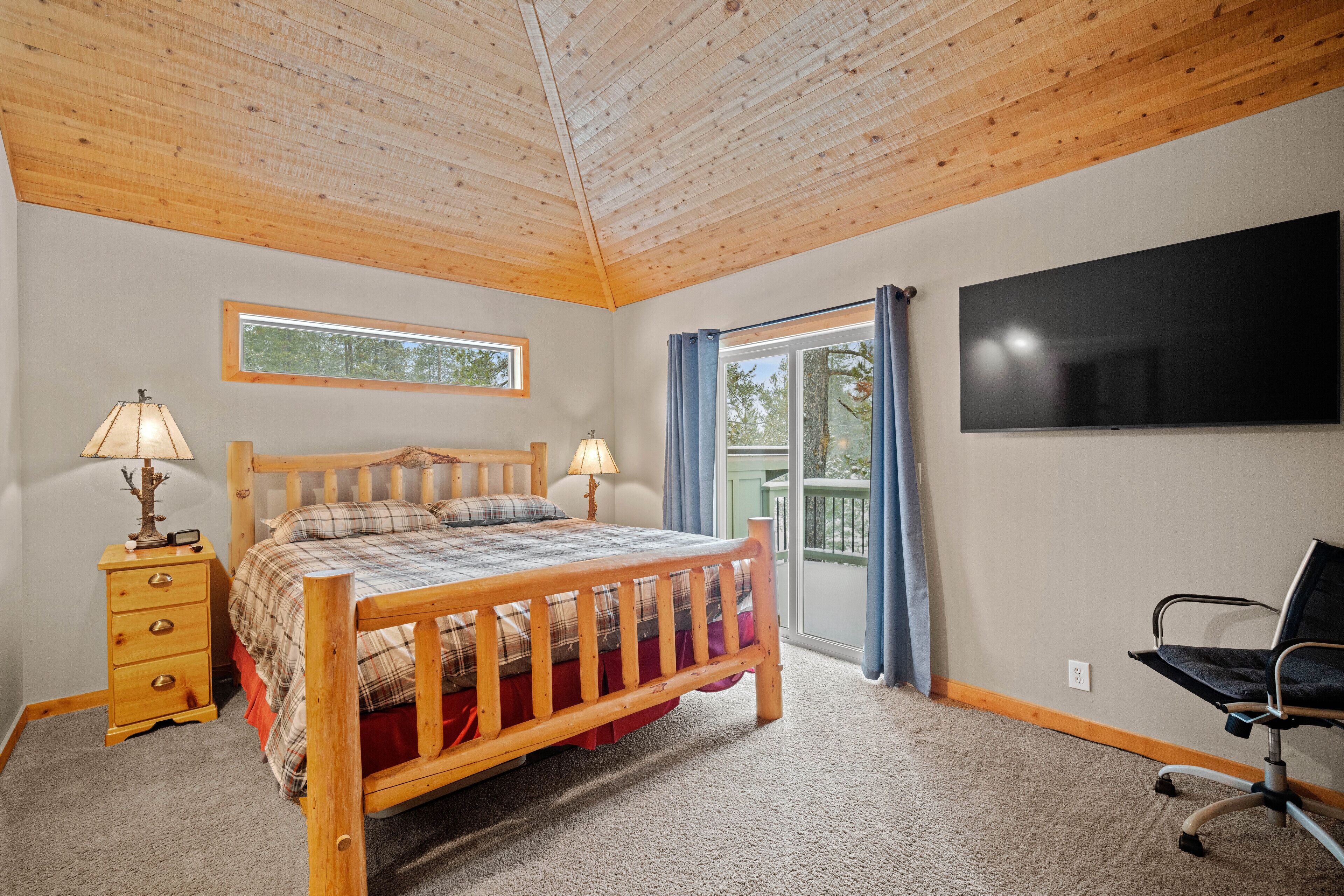 A bedroom with a wooden bed, two bedside tables with lamps, a flat-screen TV, and a view of the outdoors through a window and sliding door.