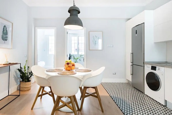 A modern kitchen with a dining area, featuring a large window, a washing machine, and a hanging light fixture.