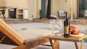 A poolside lounge area with a wooden table, two wine glasses, a bottle of wine, and a bowl of grapes.