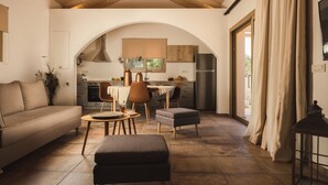 A spacious living area with a sofa, ottoman, and dining table, an arched kitchen in the background, and a large window with curtains.