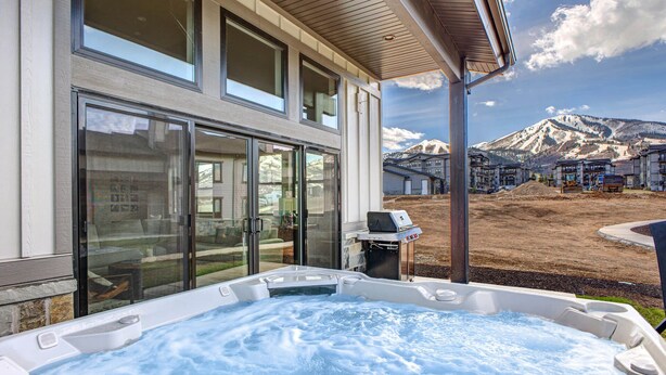 A modern building with large sliding glass doors, a hot tub, and a view of a mountain range. - Moose Mgmt at Mayflower Lakeside 1032: Central AC-Pvt Hot Tub-BBQ- Two King Beds (Park City)