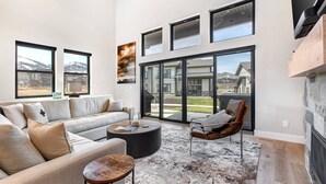 A spacious living room with a large sofa, a coffee table, and a fireplace. There are large windows and a sliding glass door leading to an outdoor area. - Moose Mgmt at Mayflower Lakeside 1032: Central AC-Pvt Hot Tub-BBQ- Two King Beds (Park City)