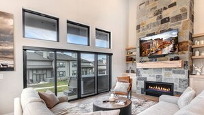 A modern living room with a stone fireplace, a large TV mounted on the wall, and a comfortable seating area with a sofa and armchairs. - Moose Mgmt at Mayflower Lakeside 1032: Central AC-Pvt Hot Tub-BBQ- Two King Beds (Park City)