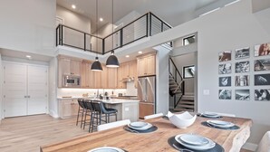 A spacious kitchen with modern appliances, a dining area with a wooden table set for four, and a staircase leading to an upper level. - Moose Mgmt at Mayflower Lakeside 1032: Central AC-Pvt Hot Tub-BBQ- Two King Beds (Park City)