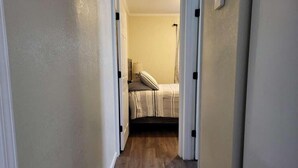 A narrow hallway with wooden flooring leading to a bedroom with a bed and a nightstand.