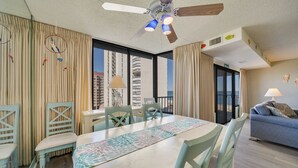 Une chambre d’hôtel comprenant un coin repas, un canapé, un ventilateur de plafond et de grandes fenêtres donnant sur la ville.