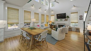 A spacious living room with a wooden dining table, wicker chairs, a fireplace, and a television mounted on the wall. - Designer Home in Seaside, FL! Walk to Pool and Beach + 2 Adult Bikes! (Santa Rosa Beach Fl)