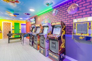 Eine hell erleuchtete Spielhalle mit mehreren Spielautomaten, darunter 'Pac-Man' und 'Mortal Kombat', und einer grünen Tür im Hintergrund.