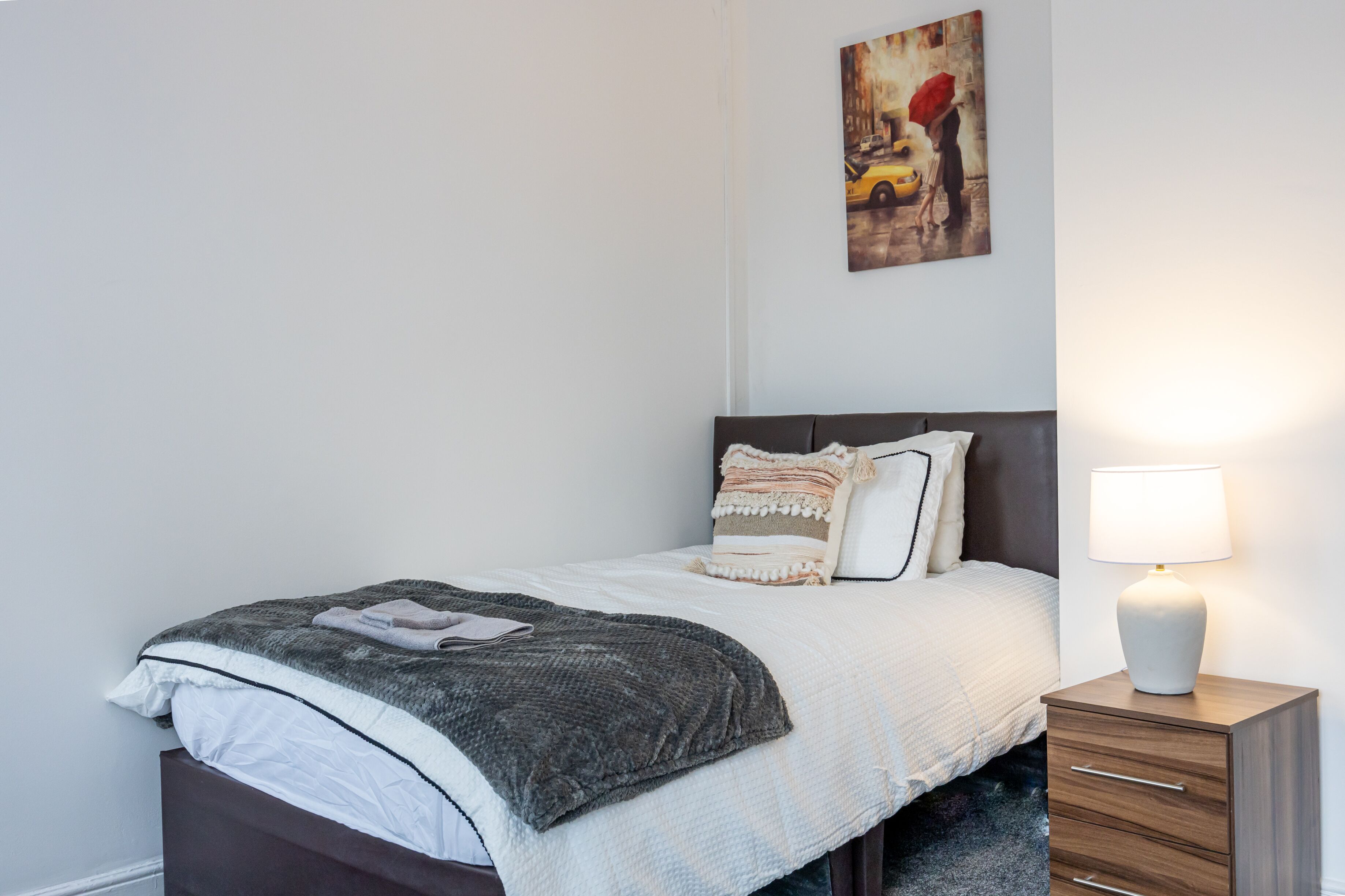 Una cama bien hecha con una colcha blanco y gris, una lámpara de noche y una mesita de madera.