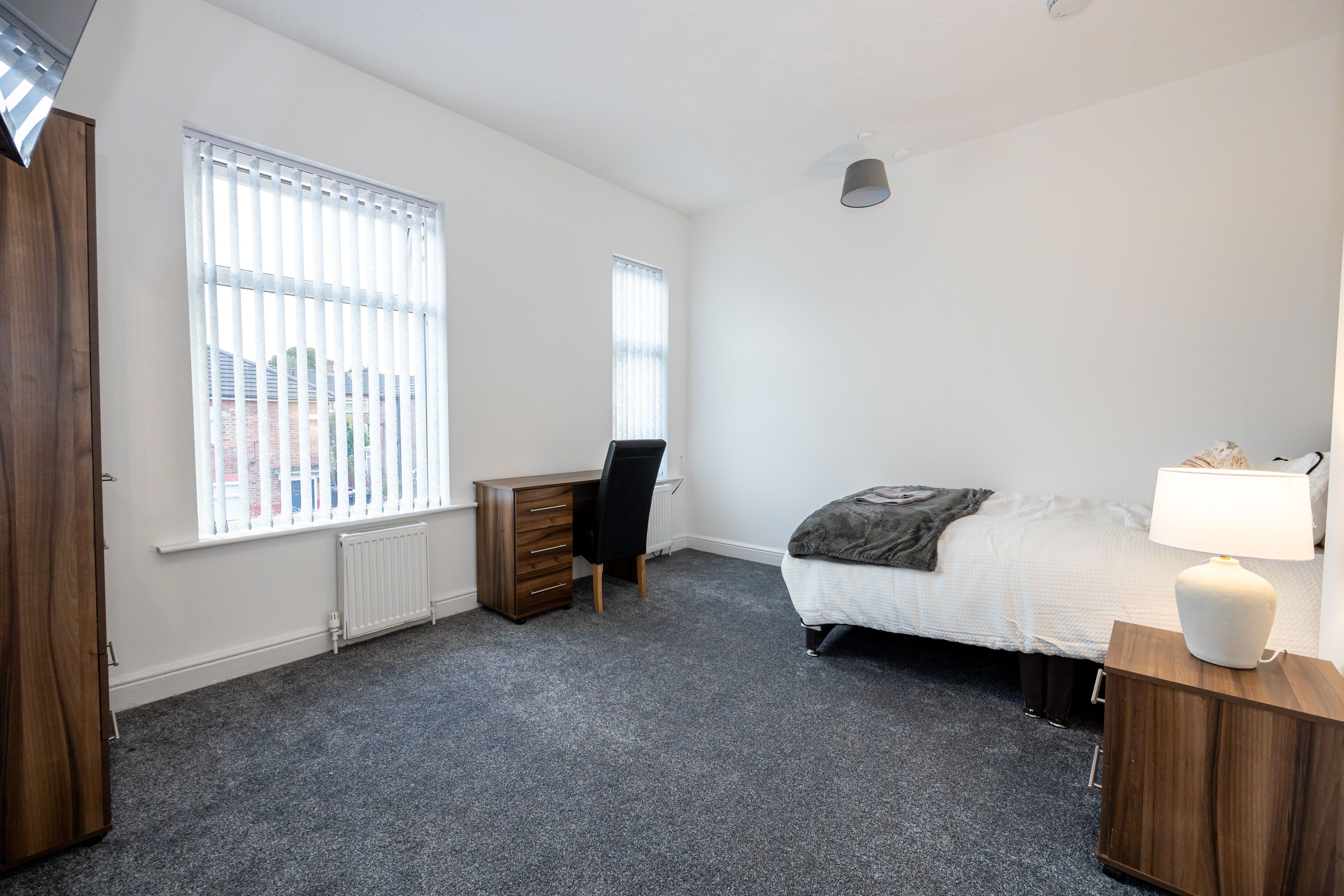 A bedroom with a bed, a desk, a chair, a radiator, and a window with blinds.