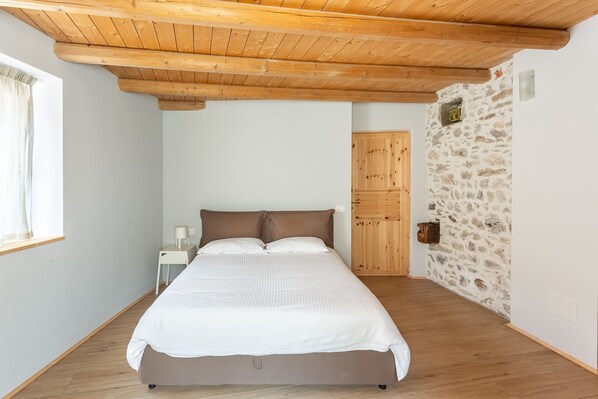 A bedroom with a wooden ceiling, a bed, a nightstand, a stone wall, and a wooden door.