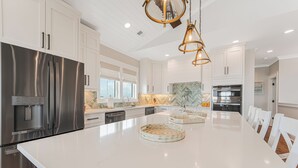 A modern kitchen with stainless steel appliances, white cabinetry, and a large island with a decorative tray. - New 8BR/7.5BA with Spectacular Ocean View, Elevator, Private Heated Pool (Ocean Isle Beach)