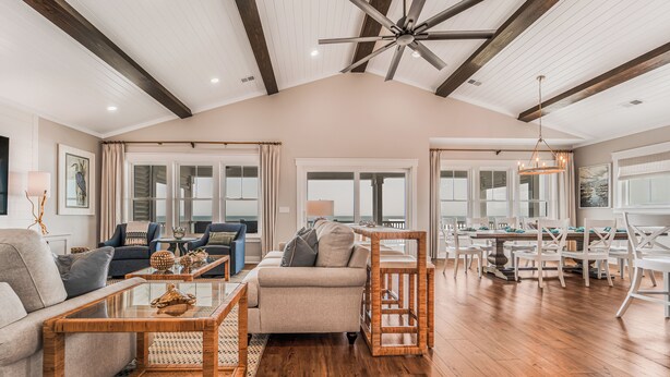 A spacious living room with a high ceiling, wooden beams, and a dining area with white chairs and a wooden table. - New 8BR/7.5BA with Spectacular Ocean View, Elevator, Private Heated Pool (Ocean Isle Beach)