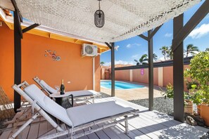A sunlit outdoor lounge area with lounge chairs, a table with a bottle and glass, and a view of a swimming pool.