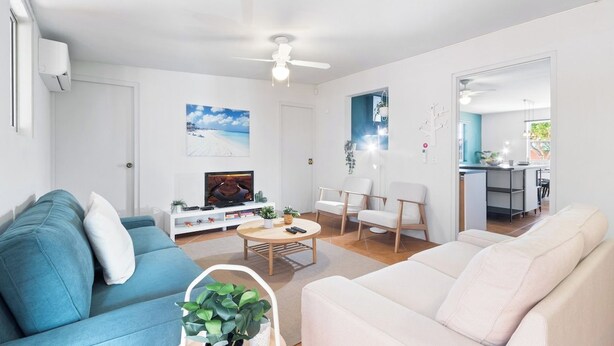Een moderne woonkamer met een blauwe bank, een witte bank, een salontafel en een televisie. Er is een plafondventilator, een schilderij en een keuken zichtbaar op de achtergrond.