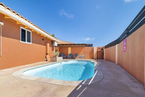 A swimming pool area with a clear blue pool, a covered seating area with chairs, and a building with a tiled roof. - Private Pool! 3BR Home w Patio near Beaches, Aruba (Noord)