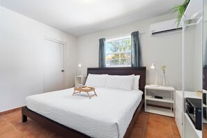 A hotel room with a bed, a bedside table with a lamp, a small shelf, and a window with curtains.