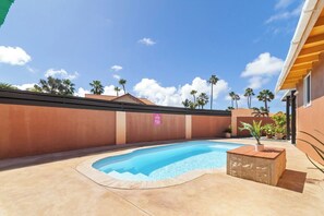 A swimming pool with a wooden deck, surrounded by a low wall and palm trees.