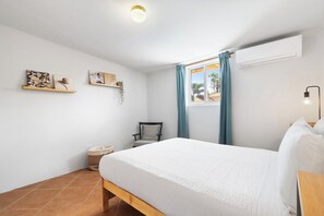 A bedroom with a bed, a chair, a window with curtains, a wall-mounted air conditioner, and artwork on the wall.