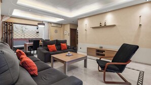 Un soggiorno moderno con divano, poltrona e tavolino. C'è anche una zona pranzo con tavolo e sedie. L'ambiente presenta una palette colori neutra e un soffitto con illuminazione a incasso.