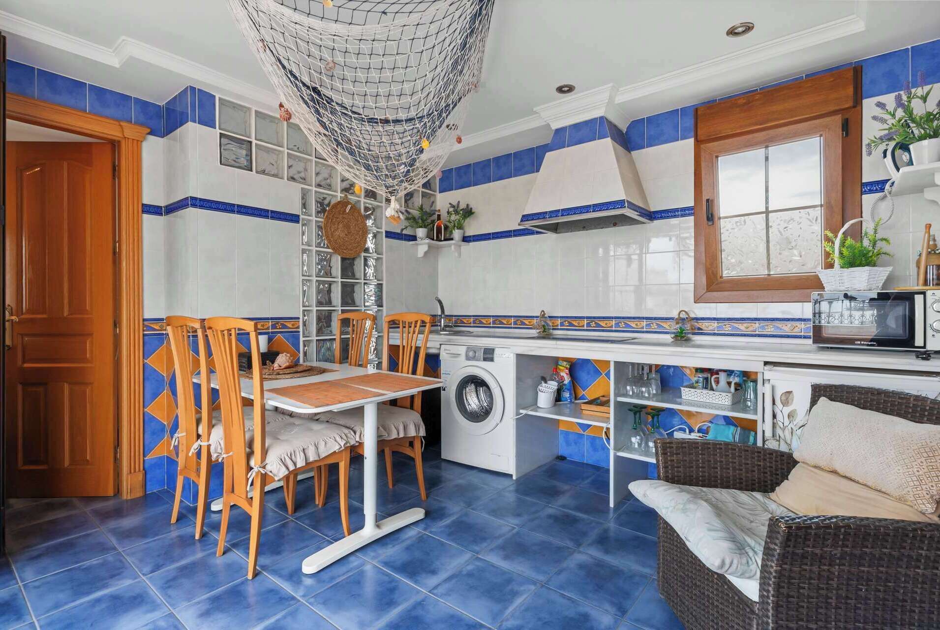 A compact kitchen with a washing machine, a dining table with chairs, and a wicker sofa.