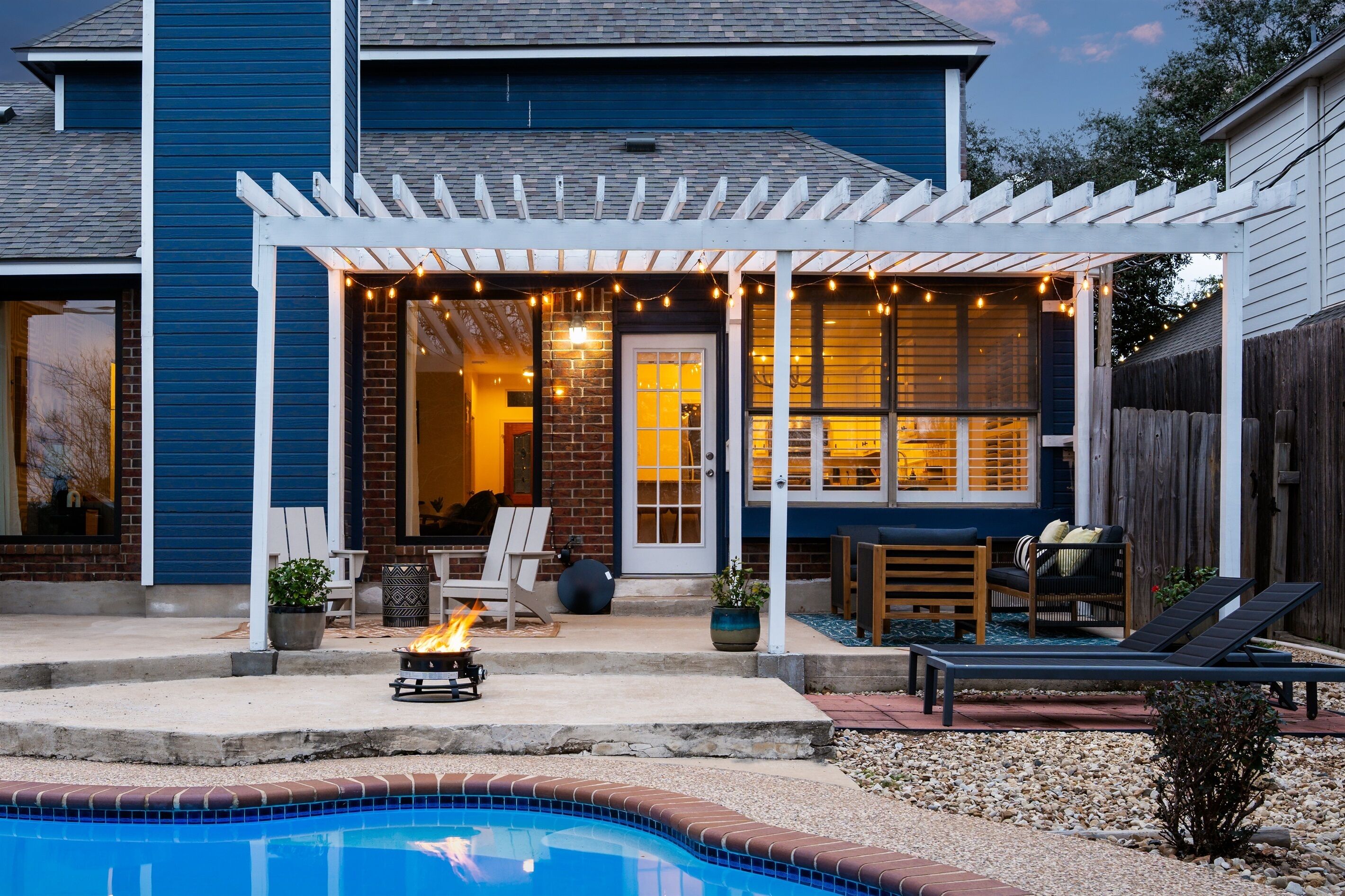 A backyard with a pool, patio seating, and a covered area with string lights.