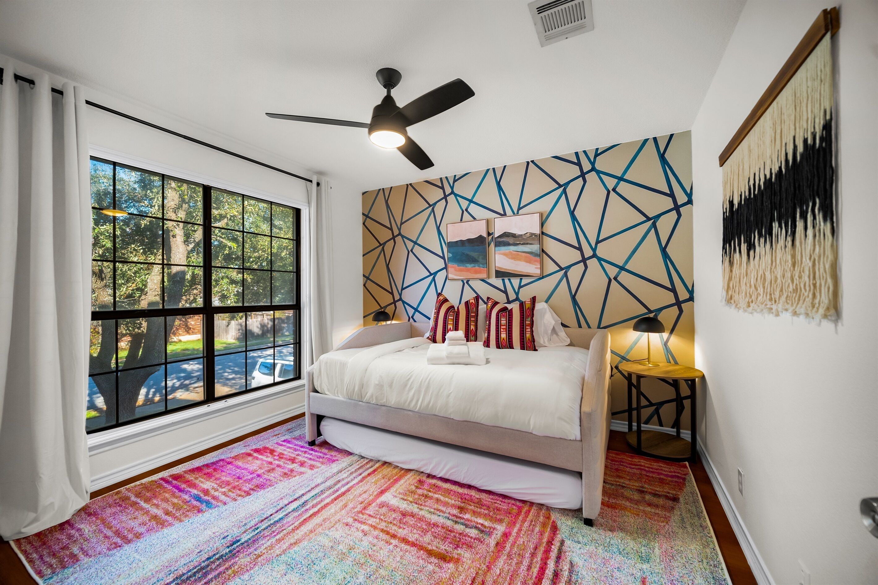 A bedroom with a large window, a ceiling fan, a bed with white bedding, a colorful rug, and a wall with geometric art.