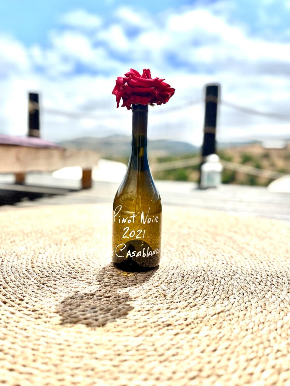 Una botella de vino Pinot Noir 2021 Casablanca sobre un tapete tejido, con un paisaje natural de fondo.