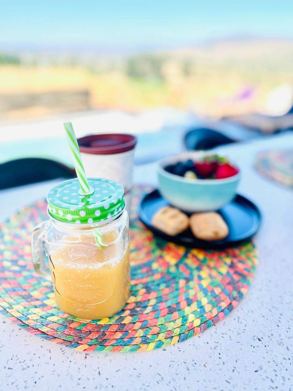 A table with a colorful woven placemat, a jar of iced tea with a green lid and straw, a bowl of berries, and a plate of cookies.