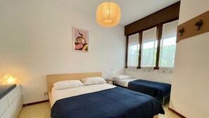 A room with two beds, a nightstand, a lamp, a window with curtains, and a wall-mounted artwork. - Spacious Duplex Near the Beach (Caorle)