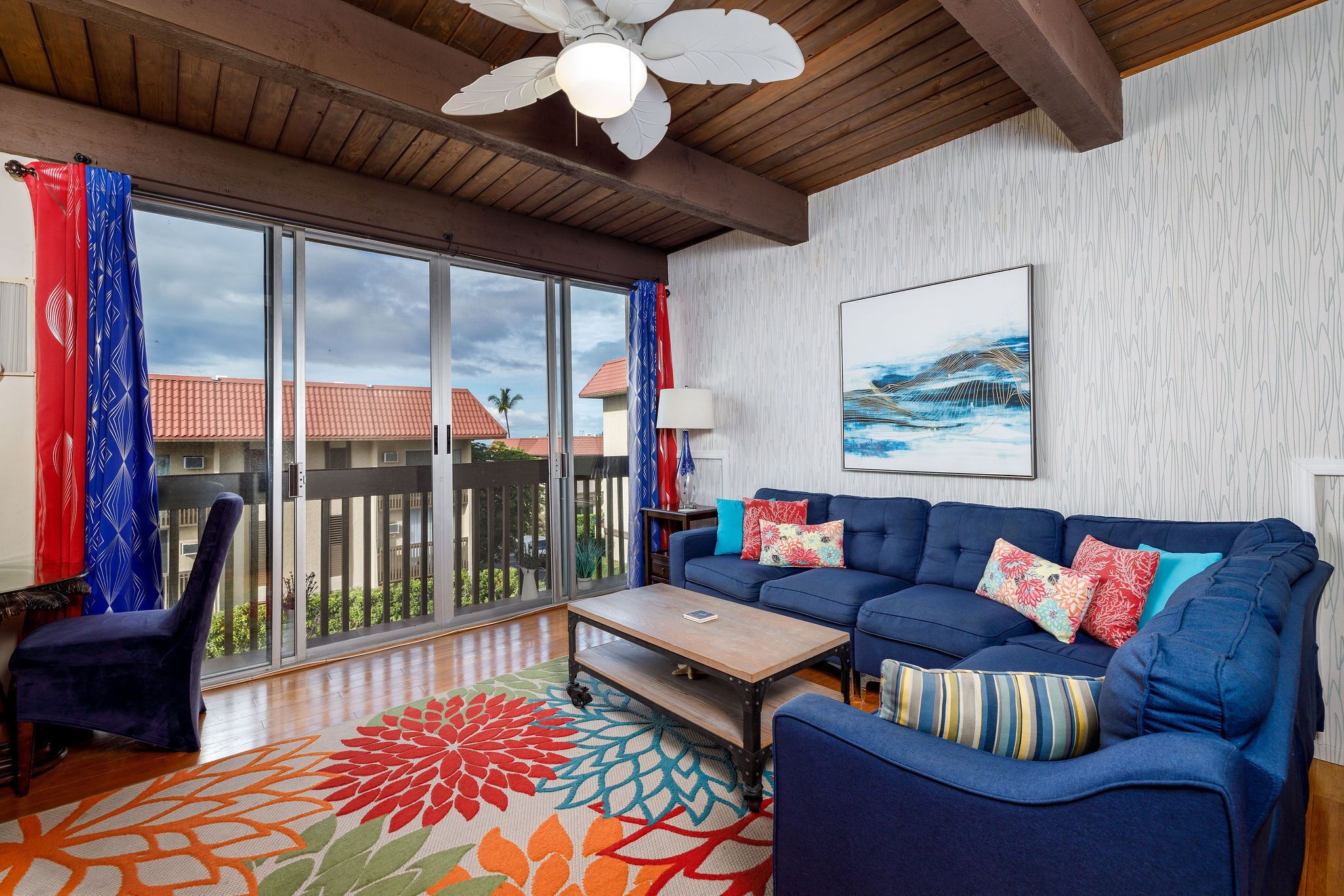 A spacious living room with a large window, a ceiling fan, a blue sofa, and a colorful rug.