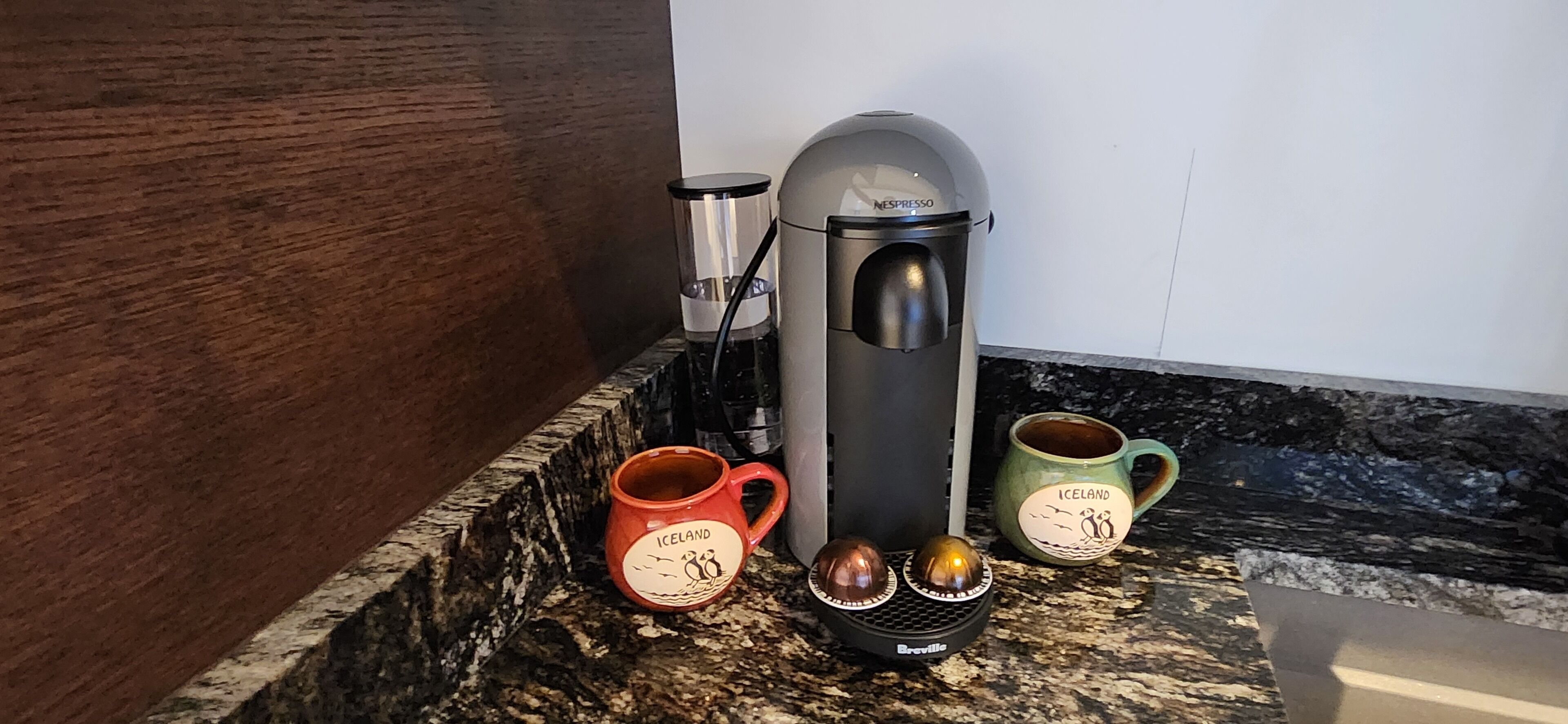 Une machine à café, deux tasses et une carafe d’eau sont posés sur un plan de travail en granit.