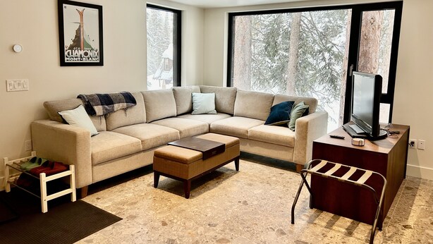 Een moderne woonkamer met een beige modulaire bank, een houten salontafel, een televisie op een standaard en een groot raam met uitzicht op bomen.