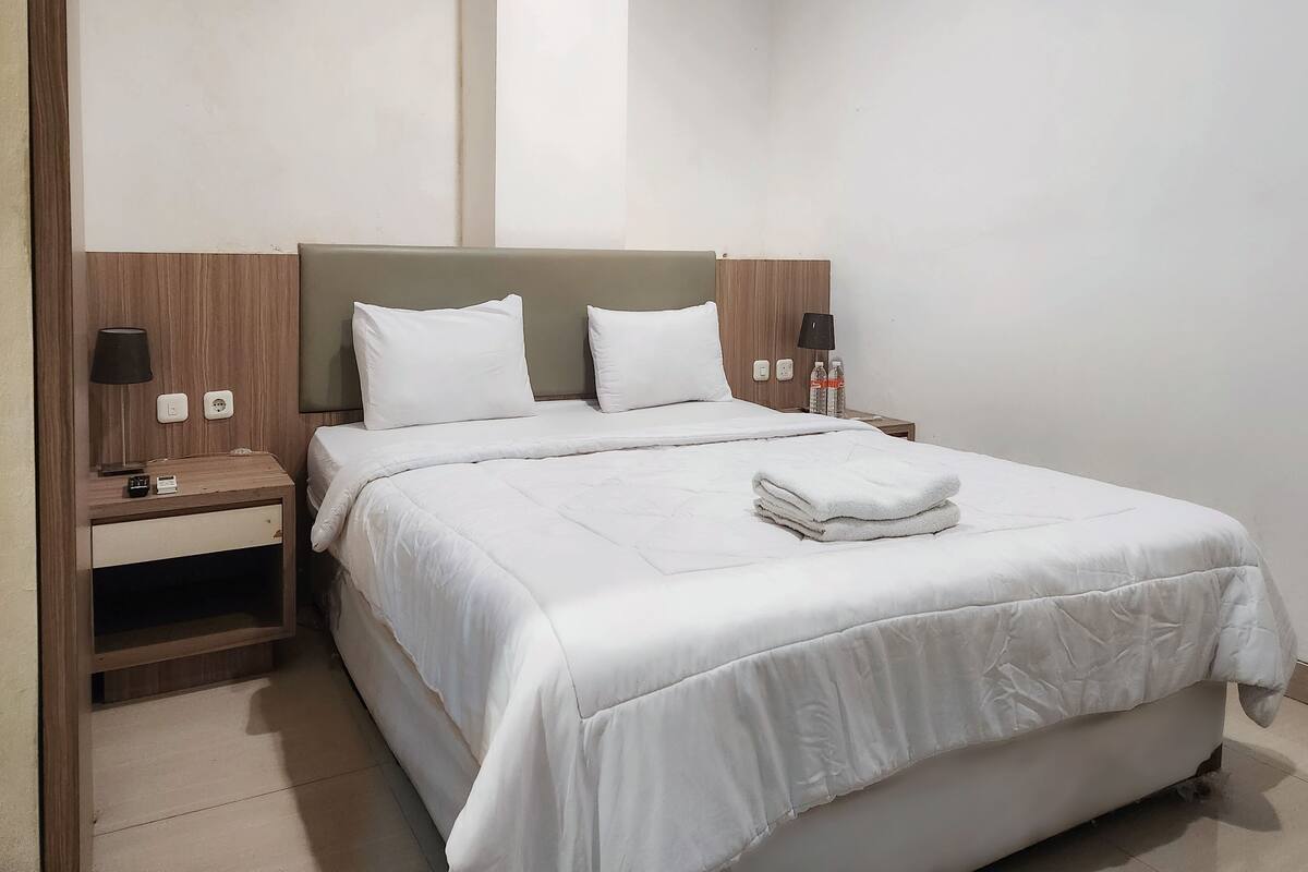 A hotel room with a large bed, two bedside tables, a nightstand, a wall-mounted TV, and a plain wall.