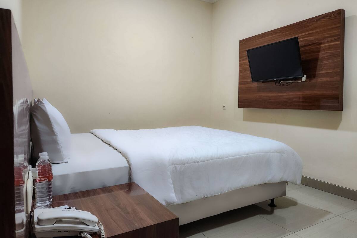 A hotel room with a bed, a wooden bedside table with a telephone, a bottle of water, and a mounted television on the wall.