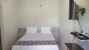 Una cama individual con una colcha blanca y estampada, dos almohadas blancas y un cabecero.  A la derecha, hay un estante pequeño con un control remoto y un enchufe.
