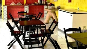 Un restaurante con paredes de ladrillo rojo, una pared de acento amarilla y un suelo de baldosas blancas. Hay varias mesas y sillas, un pequeño mostrador con un extintor, y una pizarra con el menú.