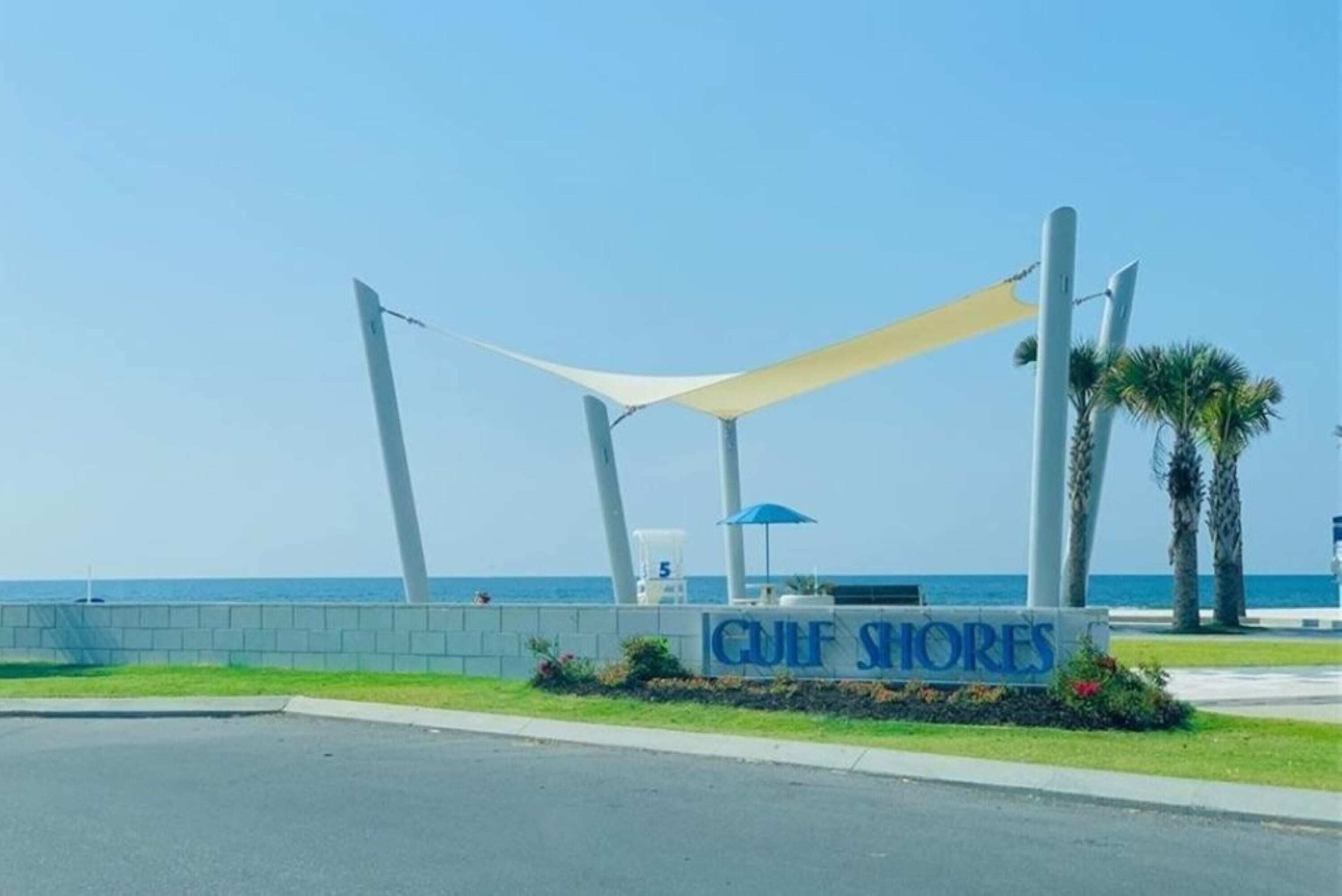A sign for 'Gulf Shores' with a beach view, a shaded seating area, and palm trees.