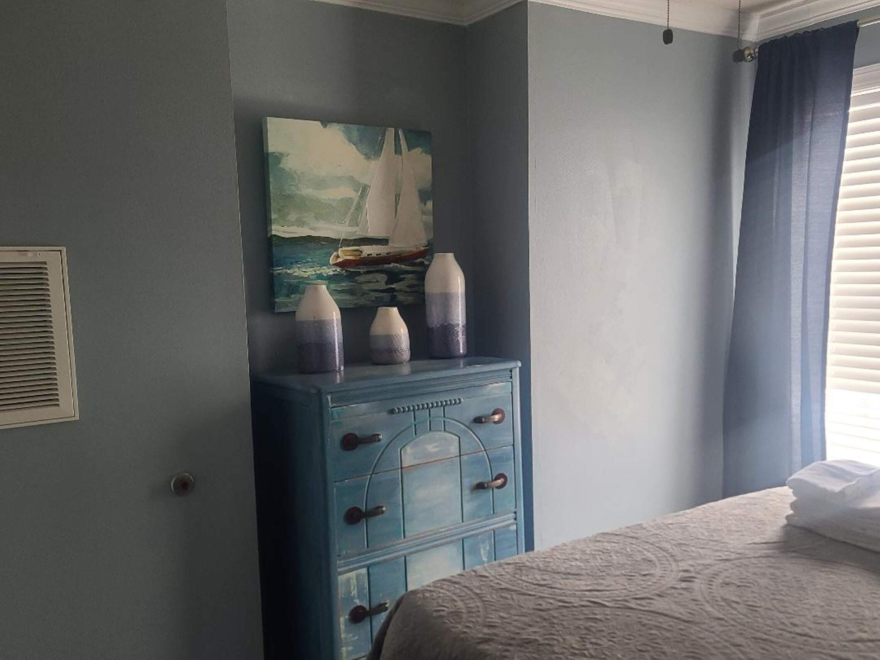 Un dormitorio con cama, cómoda con cajones, un cuadro de un velero y una ventana con cortinas.