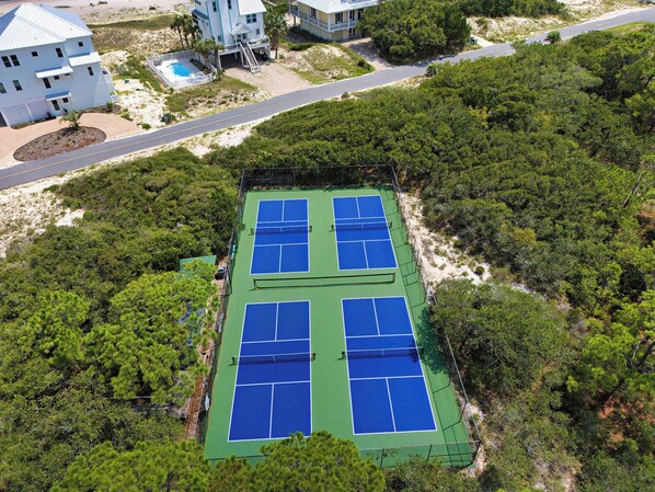 Une vue aérienne d’un court de tennis entouré d’une végétation luxuriante, avec une route et des bâtiments en arrière-plan.