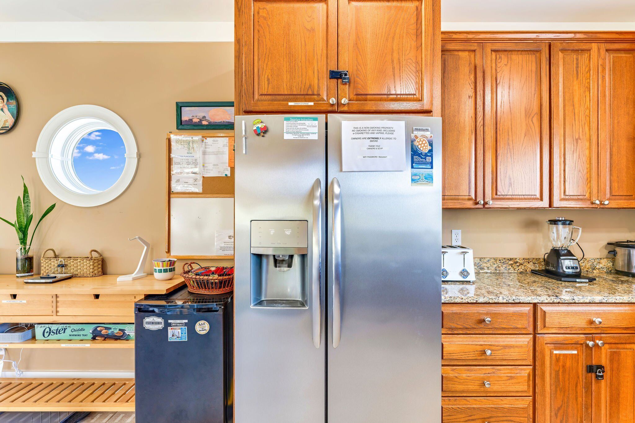 A kitchen with wooden cabinets, a stainless steel refrigerator, a water dispenser, a window with a view, and a countertop with a blender and toaster.