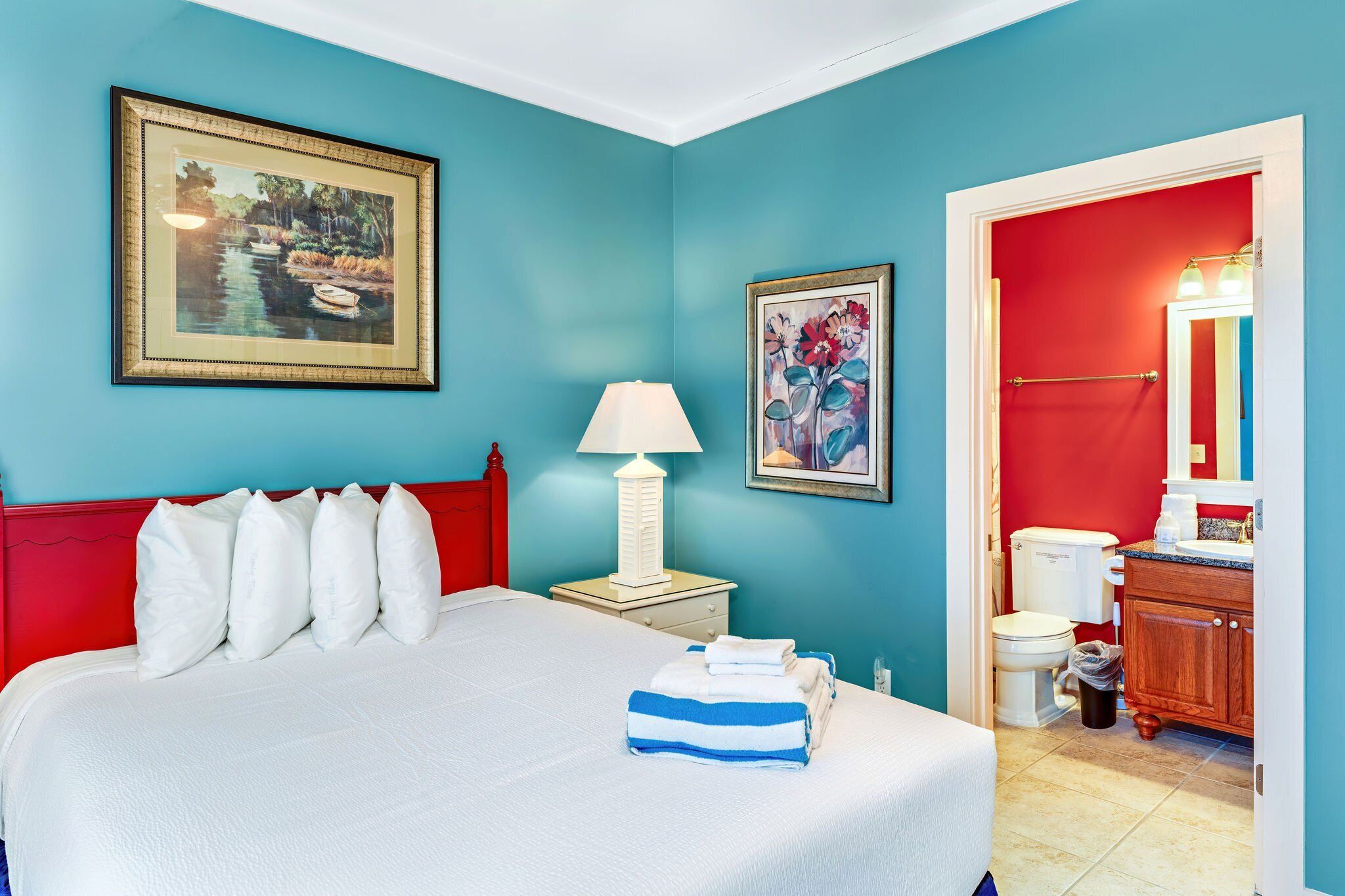 A hotel room with a bed, a nightstand with a lamp, a painting on the wall, a bathroom with a red door, and a sink.