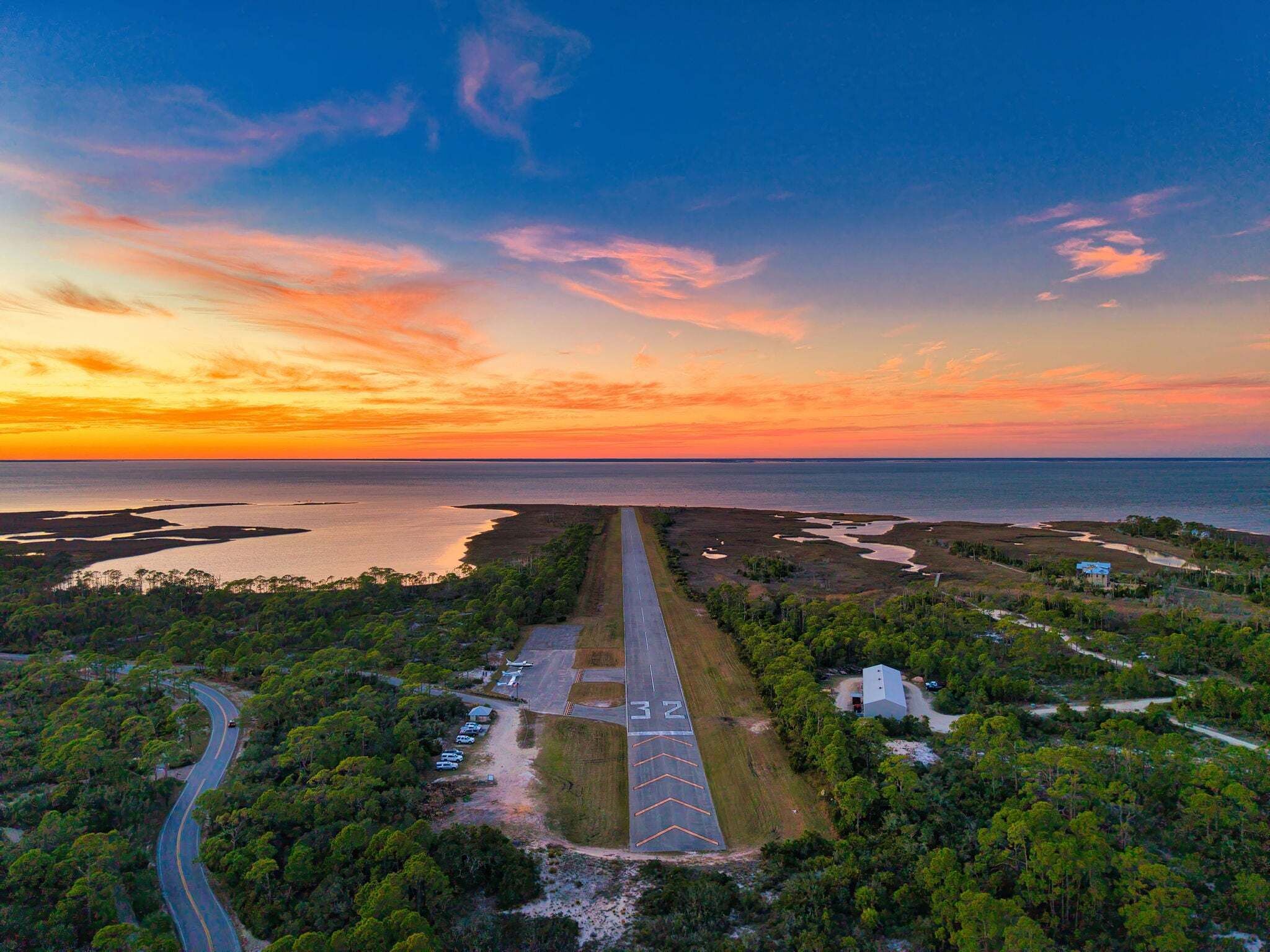 An aerial view of a coastal area with a road leading to a parking lot, surrounded by dense greenery and a body of water.